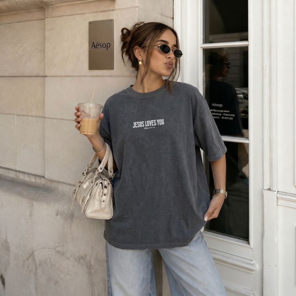 Person wearing a gray t-shirt with 'JESUS LOVES YOU' text, holding a drink and a handbag, standing outside a building.