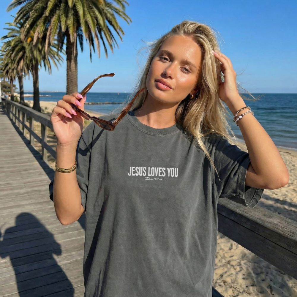 Woman wearing a 'Jesus Loves You' t-shirt on a beach with palm trees and ocean in the background.