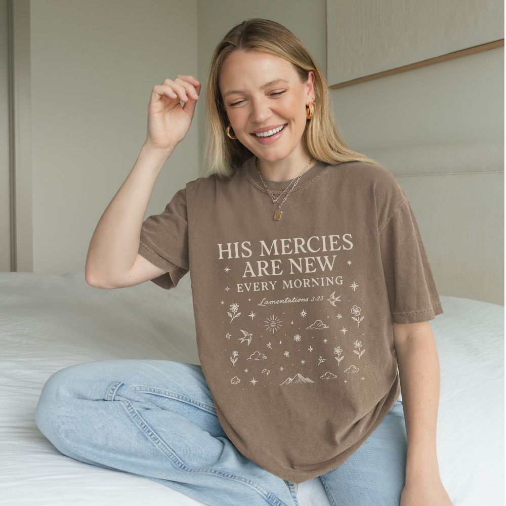 Woman wearing a brown t-shirt with 'His mercies are new every morning' text, sitting on a bed.