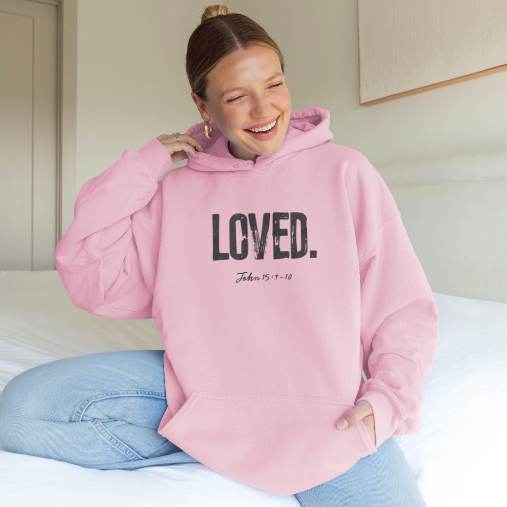 Person wearing a pink hoodie with 'LOVED' text, sitting on a bed.