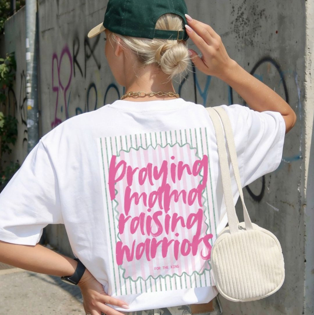 Person wearing a white t-shirt with pink text, holding a green cap, and carrying a beige bag against a graffiti-covered wall.