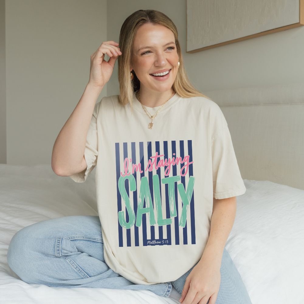 Woman wearing a t-shirt with 'I'm staying Salty' text, sitting on a bed.