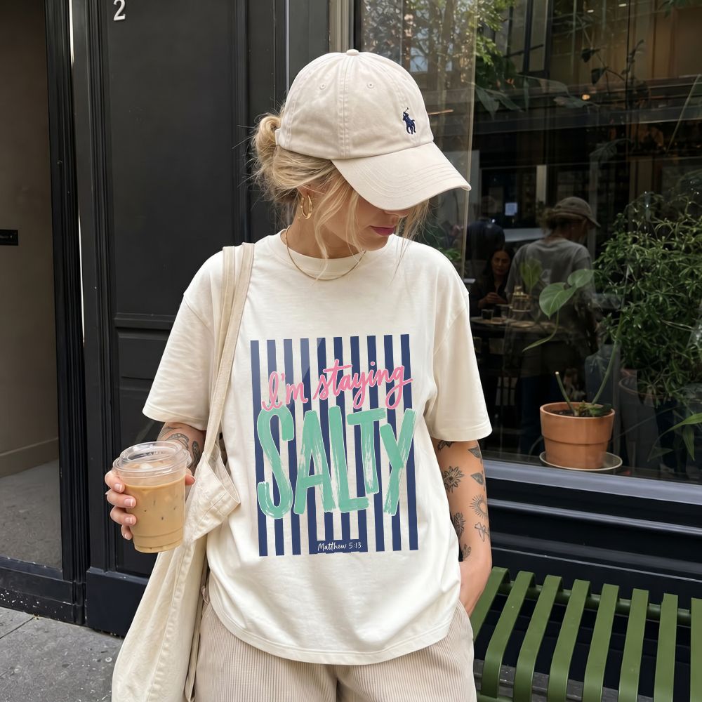 Person wearing a 'I'm staying Salty' t-shirt and beige cap, holding a drink, standing on a street.