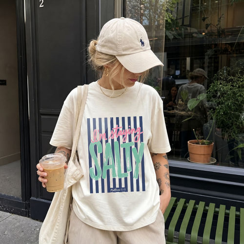 Person wearing a 'I'm staying Salty' t-shirt and beige cap, holding a drink, standing on a street.