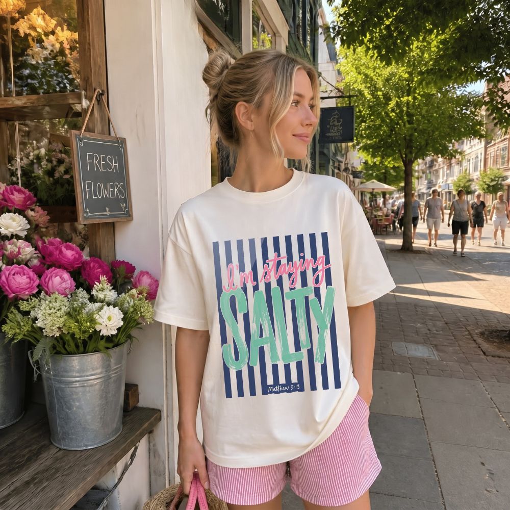 Woman wearing a 'I'm staying Salty' t-shirt on a sidewalk with flowers and a store in the background.