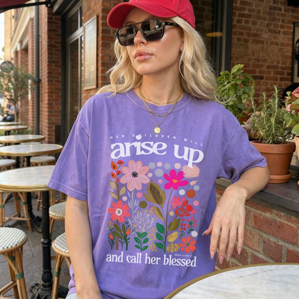 Woman wearing a purple t-shirt with floral design and text, sitting outdoors.