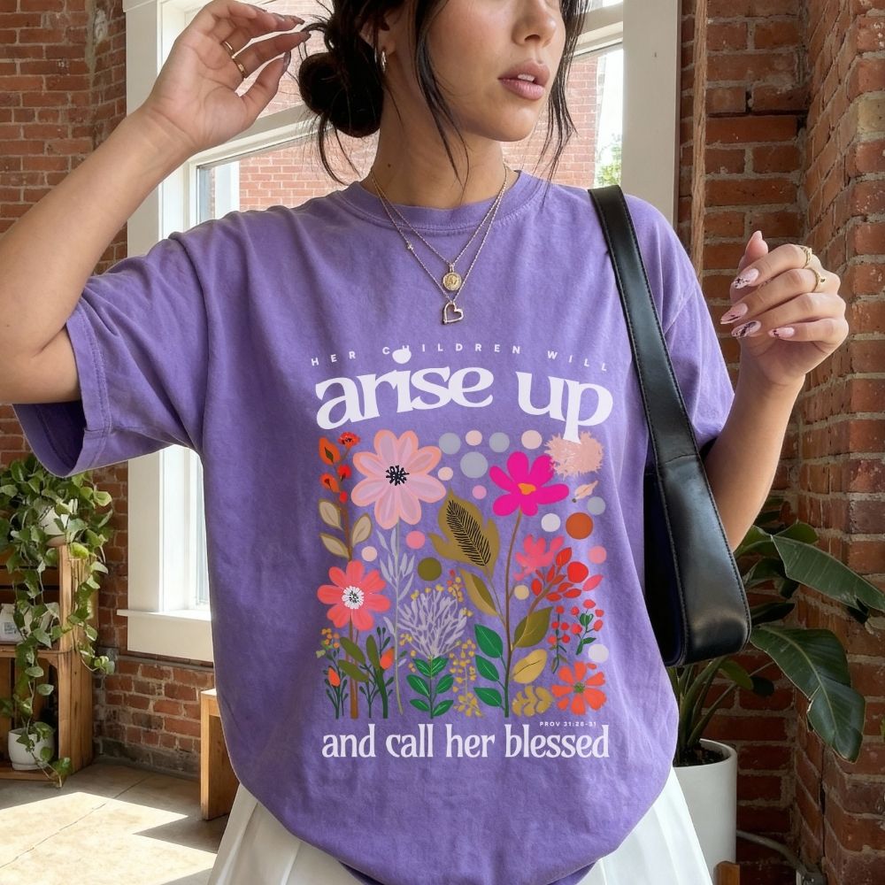 Person wearing a purple t-shirt with floral design and text in a room with brick walls and plants.