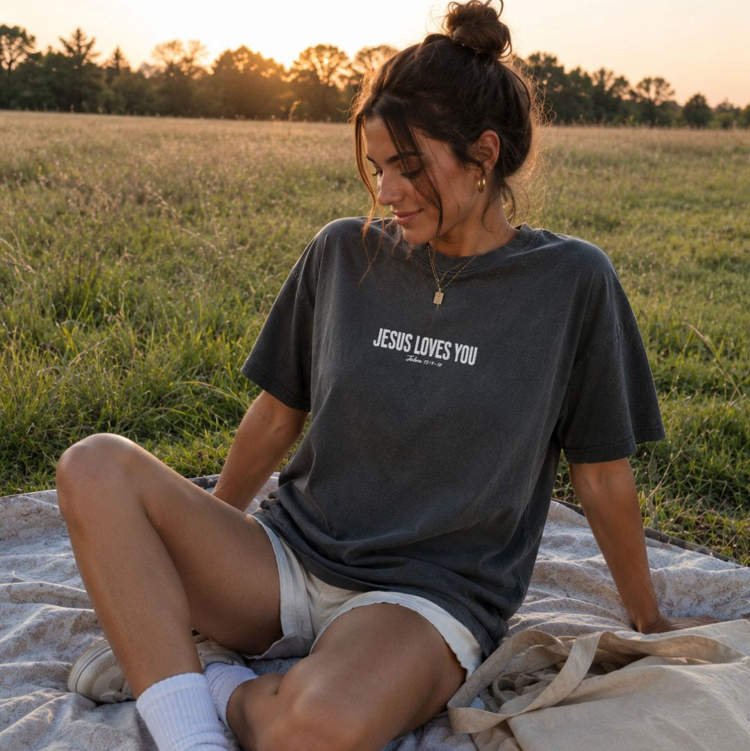 Woman sitting on a blanket in a field wearing a t-shirt with 'JESUS LOVES YOU' text.