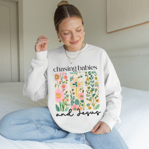 Woman wearing a white sweatshirt with floral design and text, sitting on a bed.