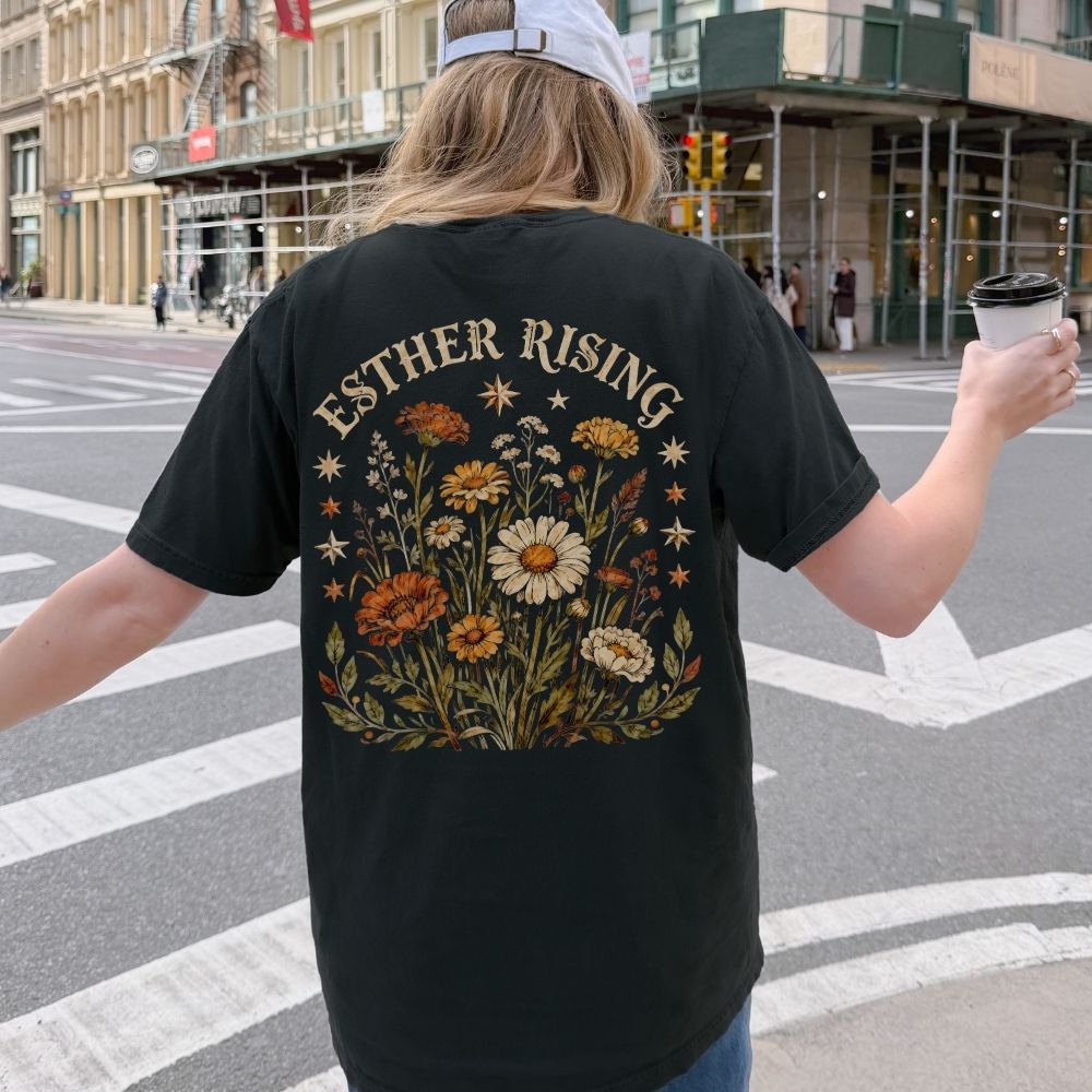 Person wearing a black t-shirt with floral design and 'Esther Rising' text on a city street.
