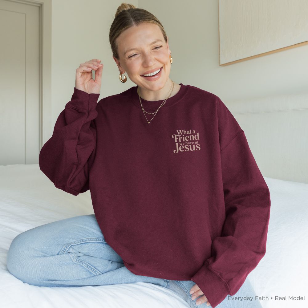 A woman sitting on a bed wearing a maroon Everyday Faith sweatshirt with the text 'What a Friend Jesus' in gold on the left chest, paired with light blue jeans. 