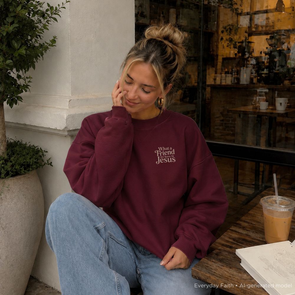 A woman sitting at a cafe table wearing a maroon Everyday Faith sweatshirt with the text 'What's Your Friend Jesus' printed in white on the left chest, paired with light blue jeans.
