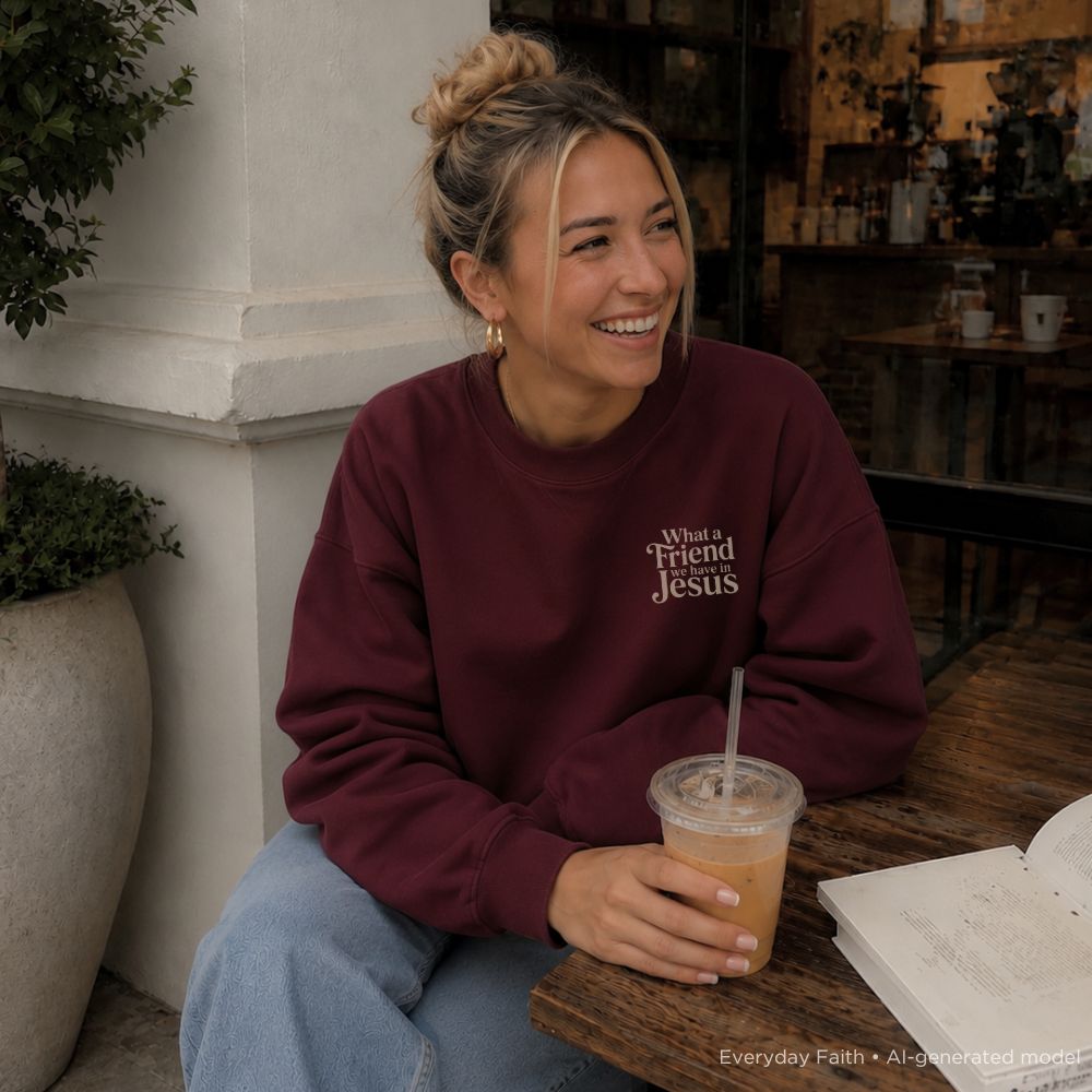 A woman sitting at a wooden table at a cafe, wearing a maroon Everyday Faith sweatshirt with the text 'What a Friend Jesus' in white. She is holding an iced coffee and wearing blue jeans.