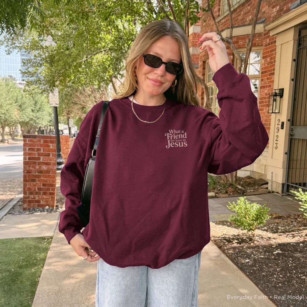 A woman wearing a maroon long-sleeve Everyday Faith sweatshirt with the text 'What's Your Friend Jesus' printed in white on the left chest, paired with light-wash jeans and sunglasses, standing outdoors on a sidewalk.