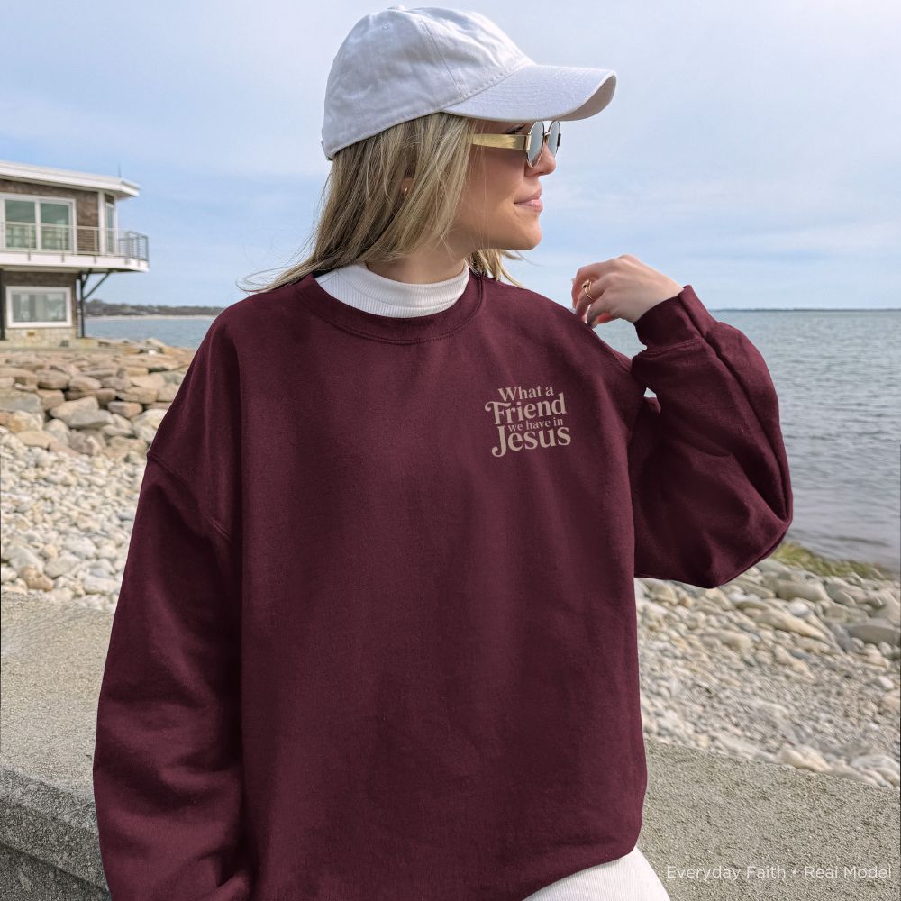 A woman wearing a maroon Everyday Faith crewneck sweatshirt with the text 'What a Friend Jesus' printed on the chest, styled with a white baseball cap, sunglasses, and a white undershirt, standing outdoors near a rocky shoreline and water.