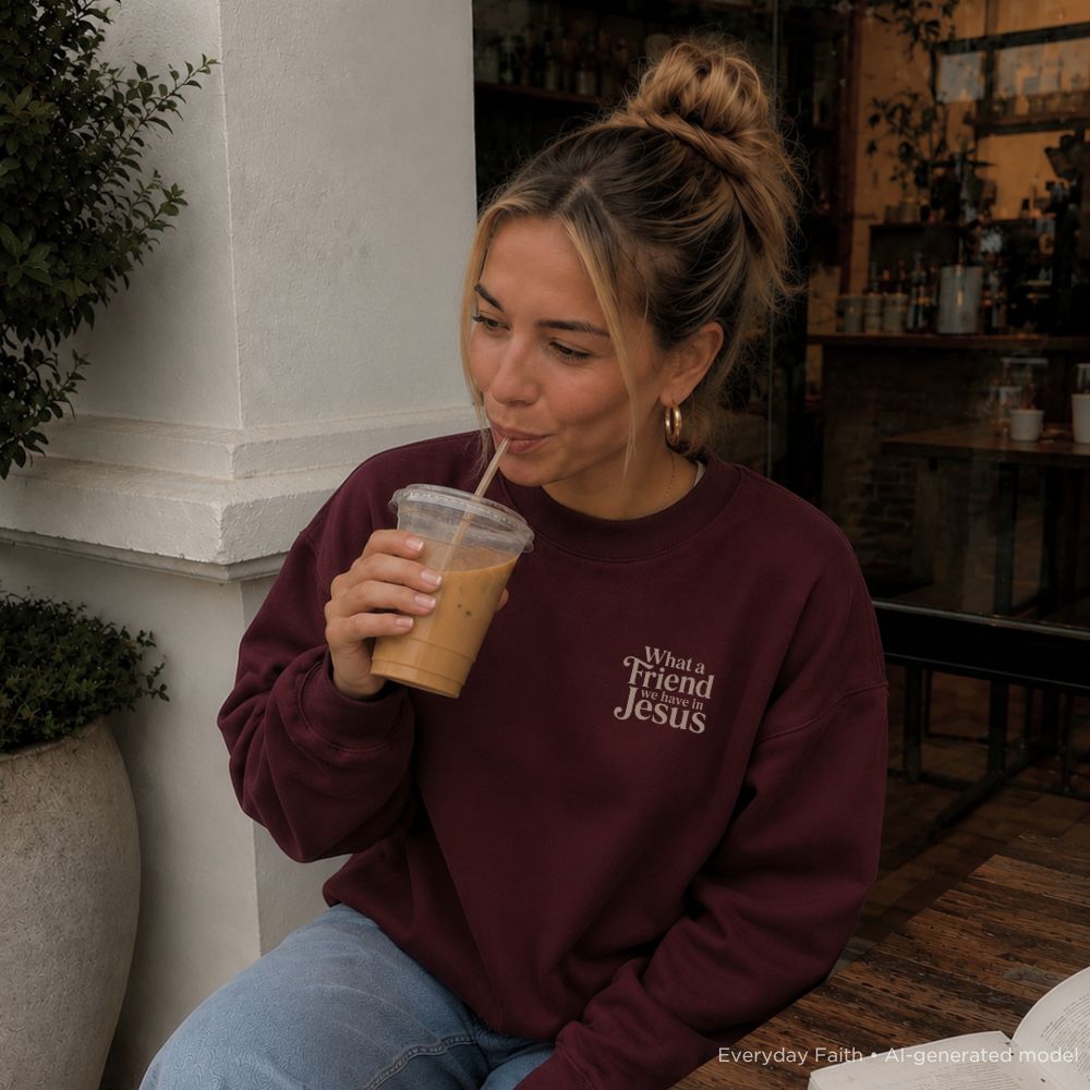 A woman with her hair in a bun sits at a cafe, drinking an iced coffee from a clear plastic cup. She is wearing a maroon Everyday Faith sweatshirt with the text 'What a friend Jesus' printed in white on the left chest, paired with light blue jeans.