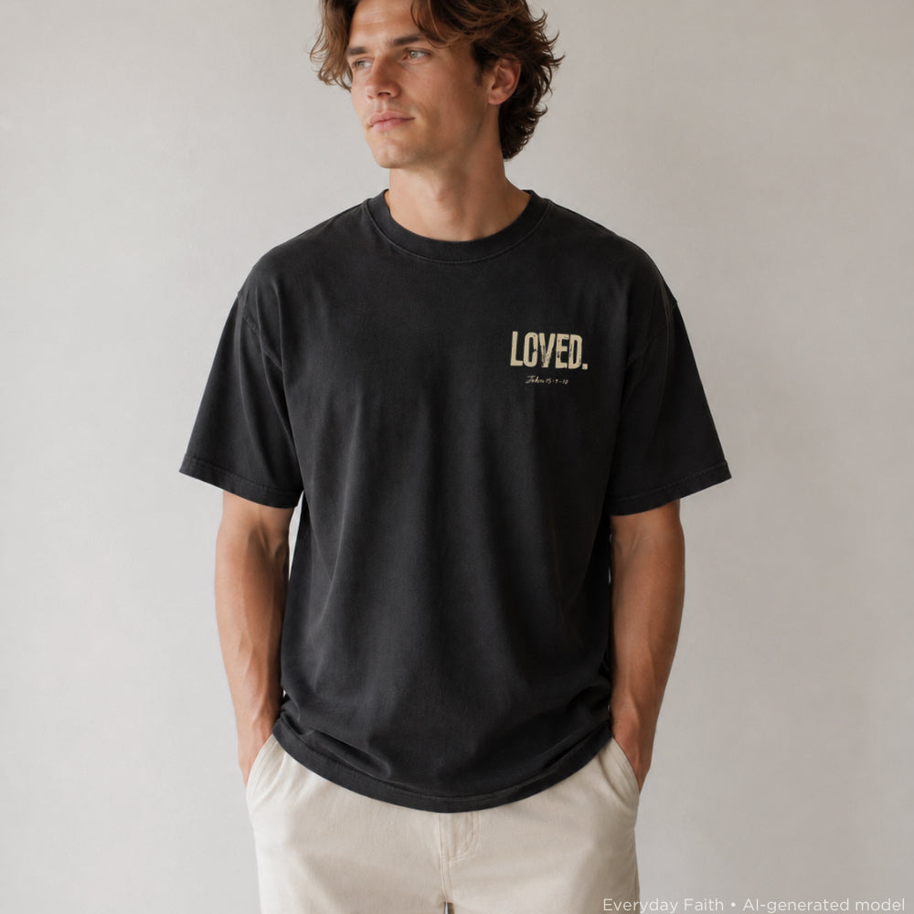 A man wearing a black Everyday Faith crewneck t-shirt with 'LOVED.' printed in beige on the left chest, paired with off-white pants, standing against a neutral light gray background.