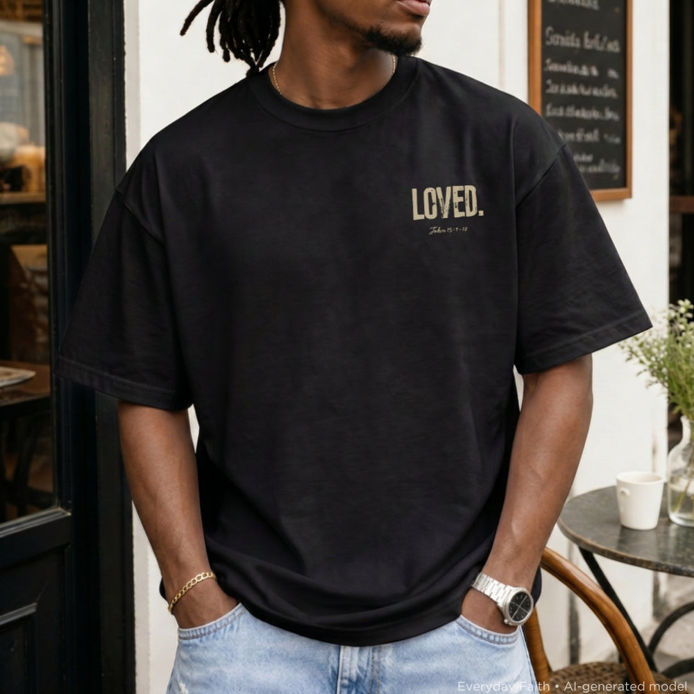 A man wearing a black Everyday Faith oversized t-shirt with 'LOVED.' printed in beige on the left chest, paired with light blue jeans, standing in front of a cafe storefront.