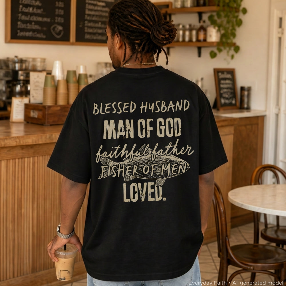 A man viewed from behind wearing a black Everyday Faith t-shirt with a humorous, religious graphic printed on the back in a cafe setting. The text reads: 'BLESSED HUSBAND MAN OF GOD faithful father FISHER OF MEN LOVED.