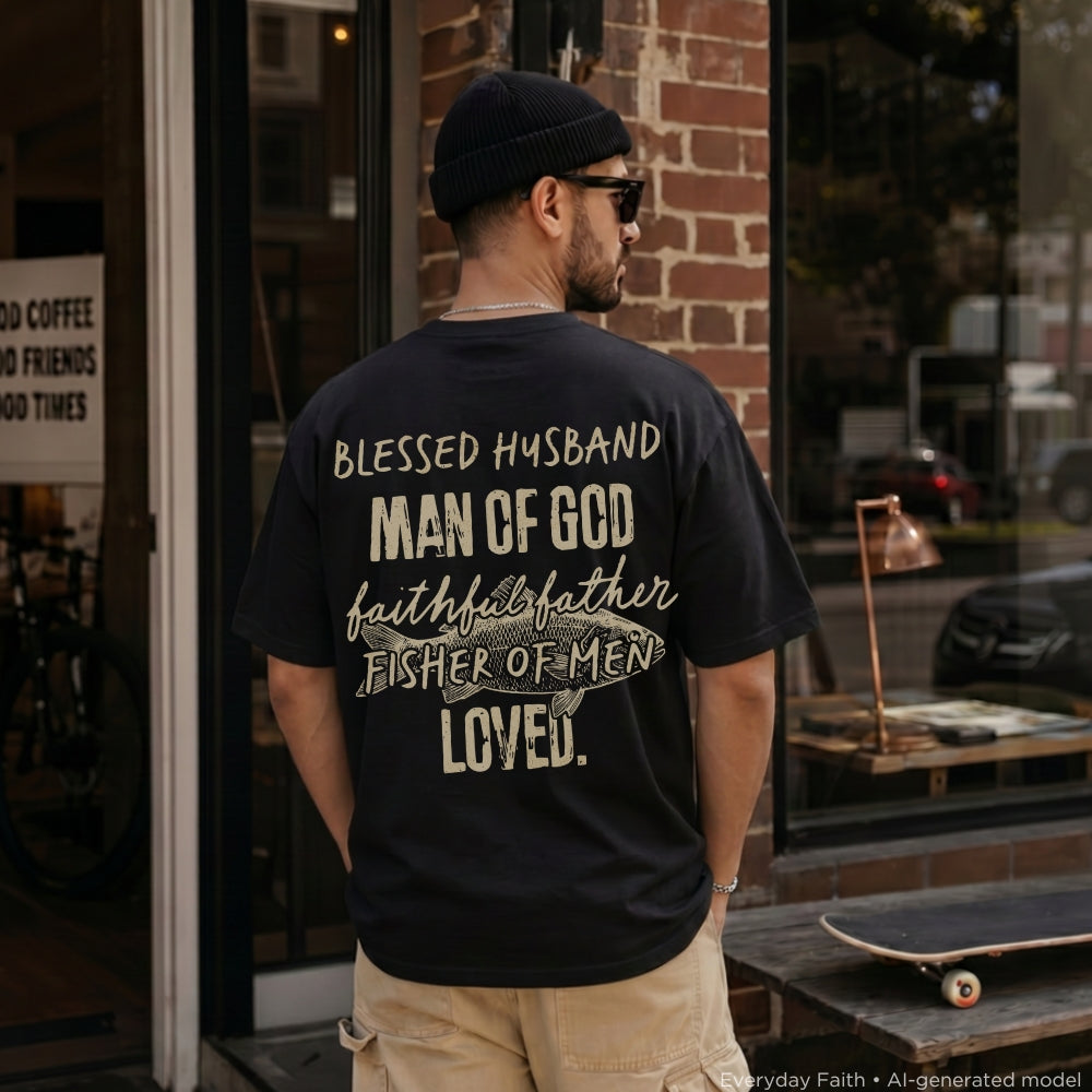 A man wearing a black Everyday Faith t-shirt with a large white and beige graphic text on the back, standing in front of a brick shop window.
