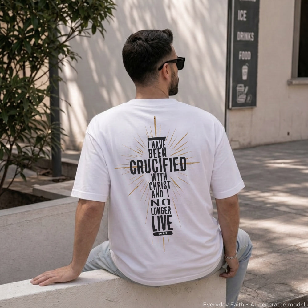 A man wearing a white Everyday Faith graphic t-shirt sits on a concrete ledge outdoors, viewed from behind. The back of the t-shirt features a black typographic design with a cross and sunburst graphic that reads 'I HAVE BEEN CRUCIFIED WITH CHRIST AND NO LONGER LIVE' in all caps.