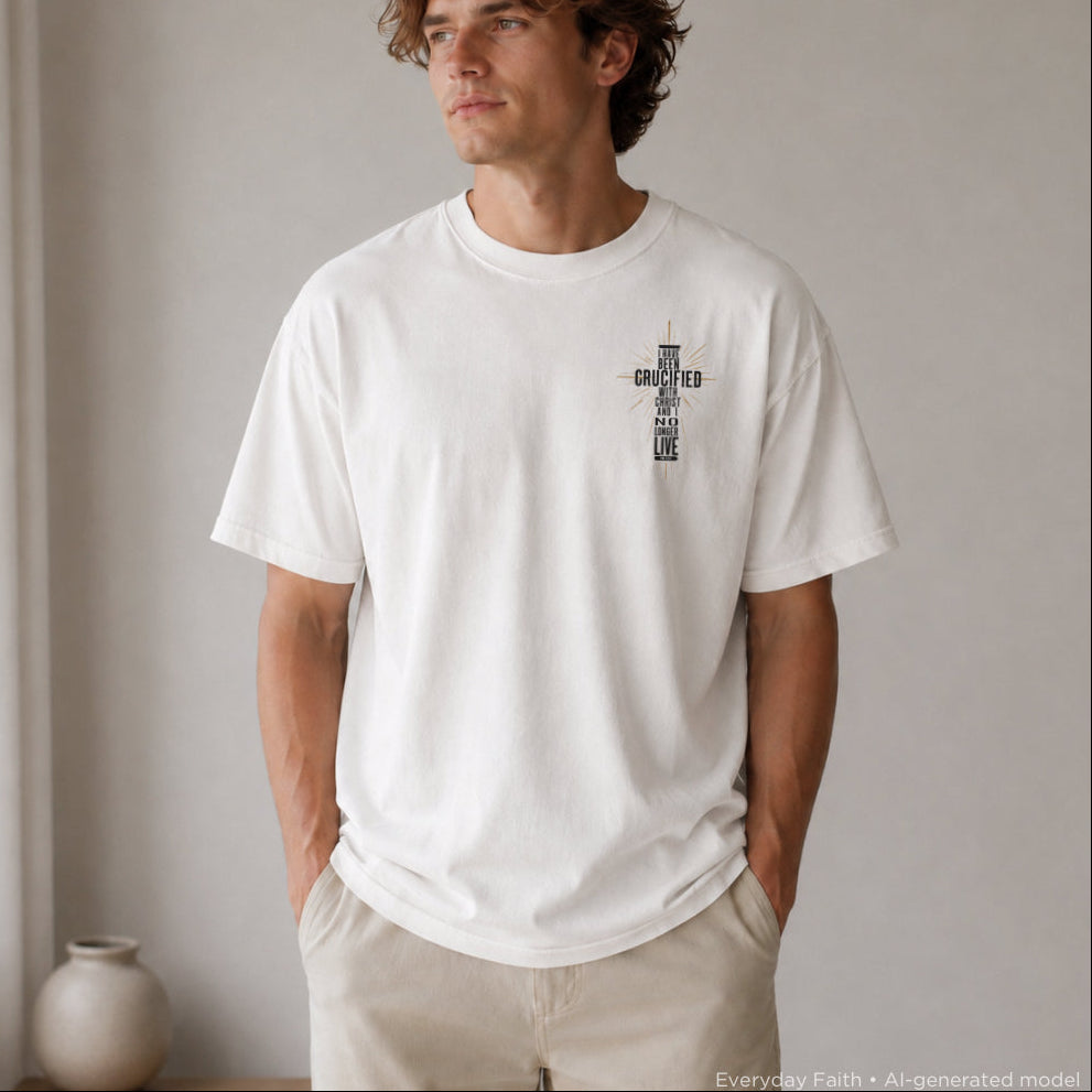 A man wearing a white Everyday Faith crewneck t-shirt with a black cross graphic on the chest containing the words 'CRUCIFIED WITH CHRIST' in vertical text, paired with beige pants in a minimalist indoor setting.