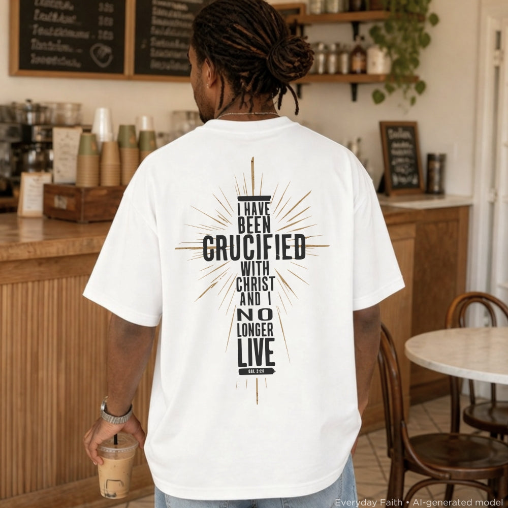 A man viewed from behind wearing a white Everyday Faith graphic t-shirt with a black and gold text design that reads 'I HAVE BEEN CRUCIFIED WITH CHRIST AND I NO LONGER LIVE FOR MYSELF'. The setting is a warm, wood-paneled cafe interior.