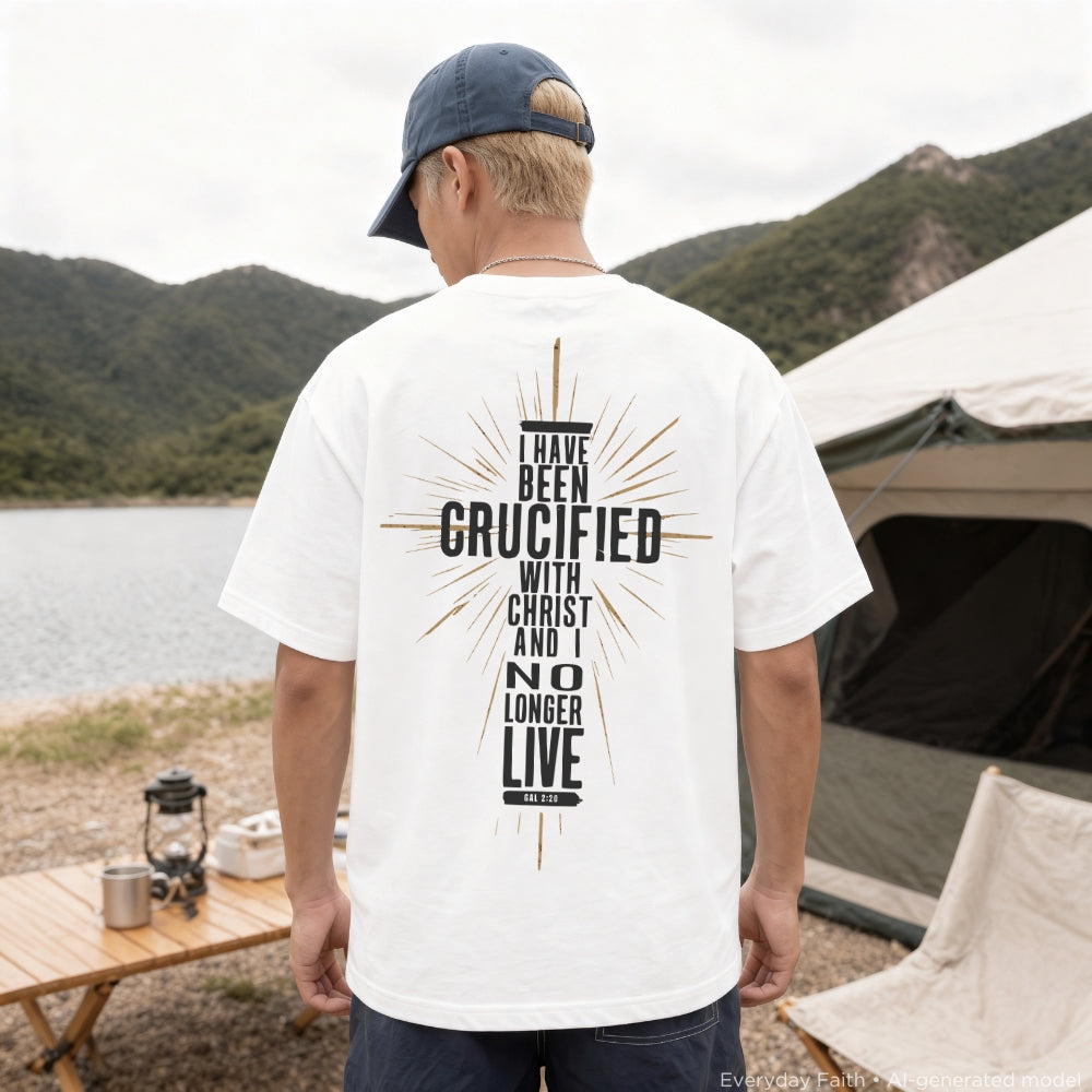 A man standing outdoors at a campsite with a lake and mountains in the background, wearing a white Everyday Faith t-shirt with a large black and gold graphic print on the back that reads 'I HAVE BEEN CRUCIFIED WITH CHRIST AND I NO LONGER LIVE ON MY OWN'.