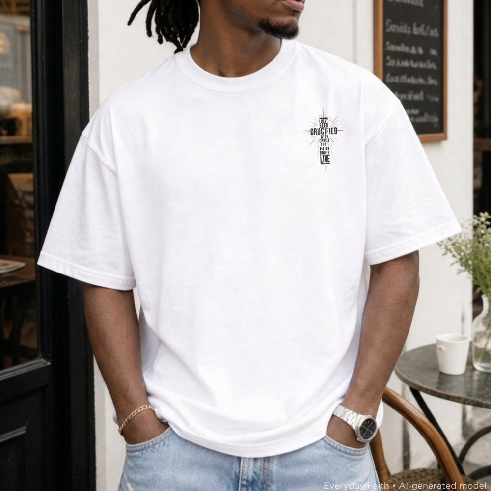 A man wearing a white Everyday Faith crewneck t-shirt with the black handwritten text 'I HAVE BEEN CRUCIFIED WITH CHRIST' on the left chest, paired with light blue jeans, standing in front of a cafe doorway.