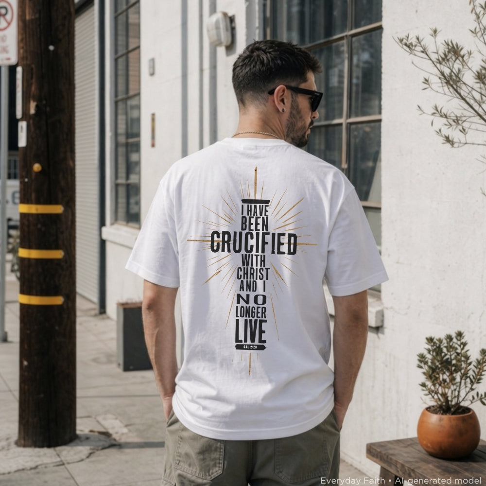 A man viewed from behind wearing a white Everyday Faith graphic t-shirt with a black and gold religious design that reads 'I HAVE BEEN CRUCIFIED WITH CHRIST'. He is standing outdoors on a city sidewalk next to a white building.