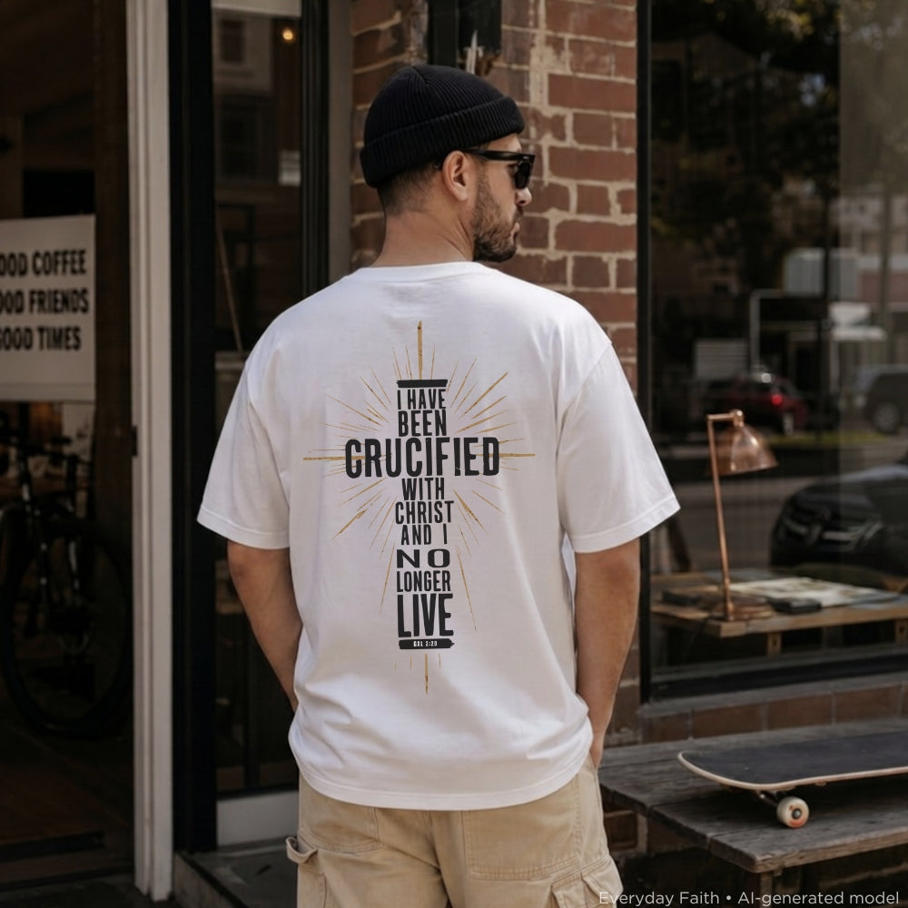 A man wearing a white Everyday Faith graphic t-shirt with a black and gold 'I Have Been Crucified With Christ and No Longer Live' print on the back, standing in front of a brick storefront.