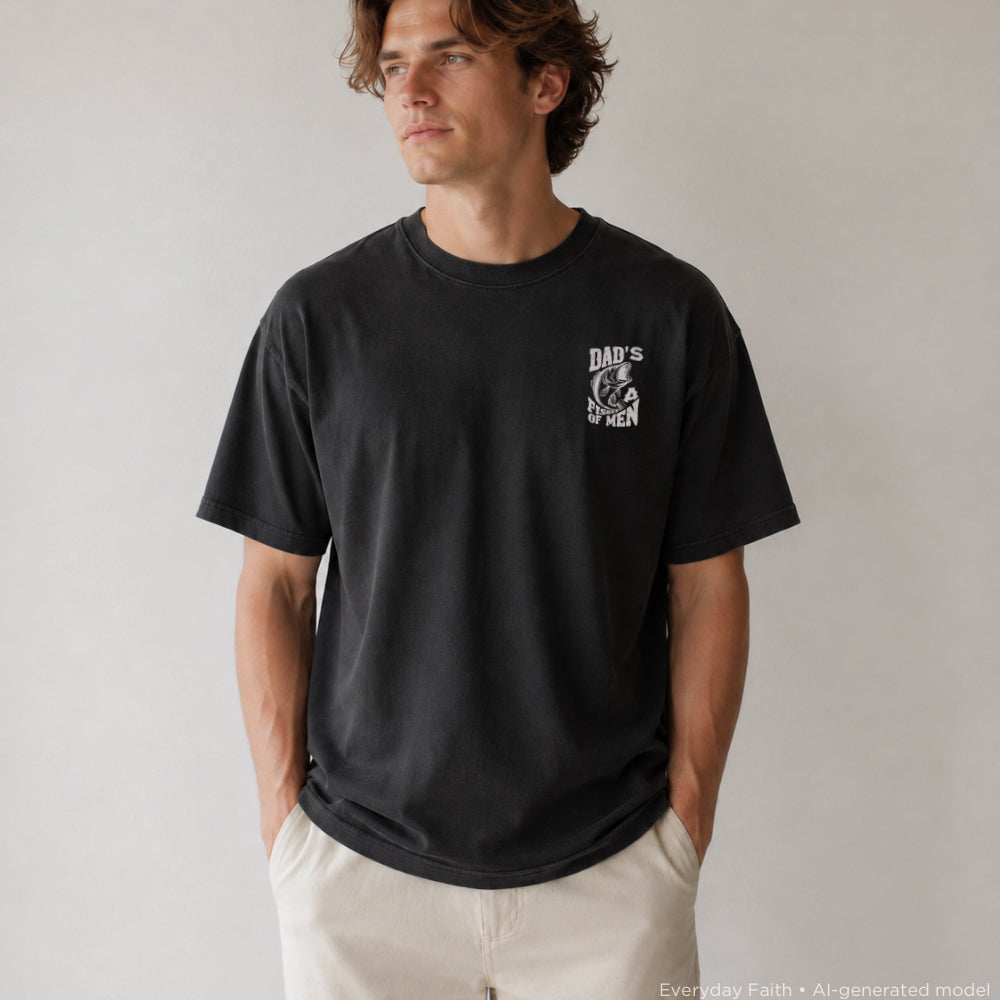 A man wearing a black Everyday Faith crewneck t-shirt with a white graphic on the left chest that reads 'DAD'S A FISHER OF MEN' with a hand holding a lightbulb, paired with off-white pants, set against a plain light gray background.