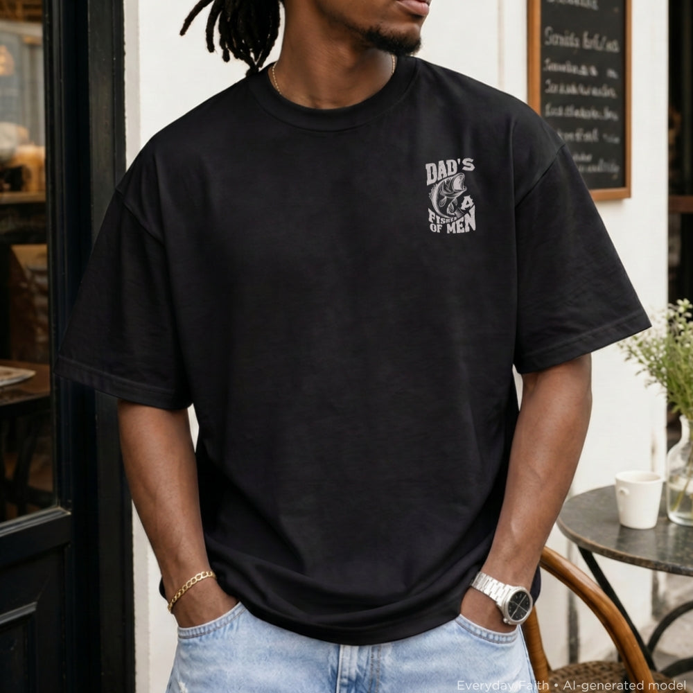 A man wearing a black oversized Everyday Faith t-shirt with a distressed white graphic on the left chest that reads 'DAD'S OF MEN' with a silhouette of a man holding a beer glass. He is wearing light blue jeans and standing in front of a cafe storefront.