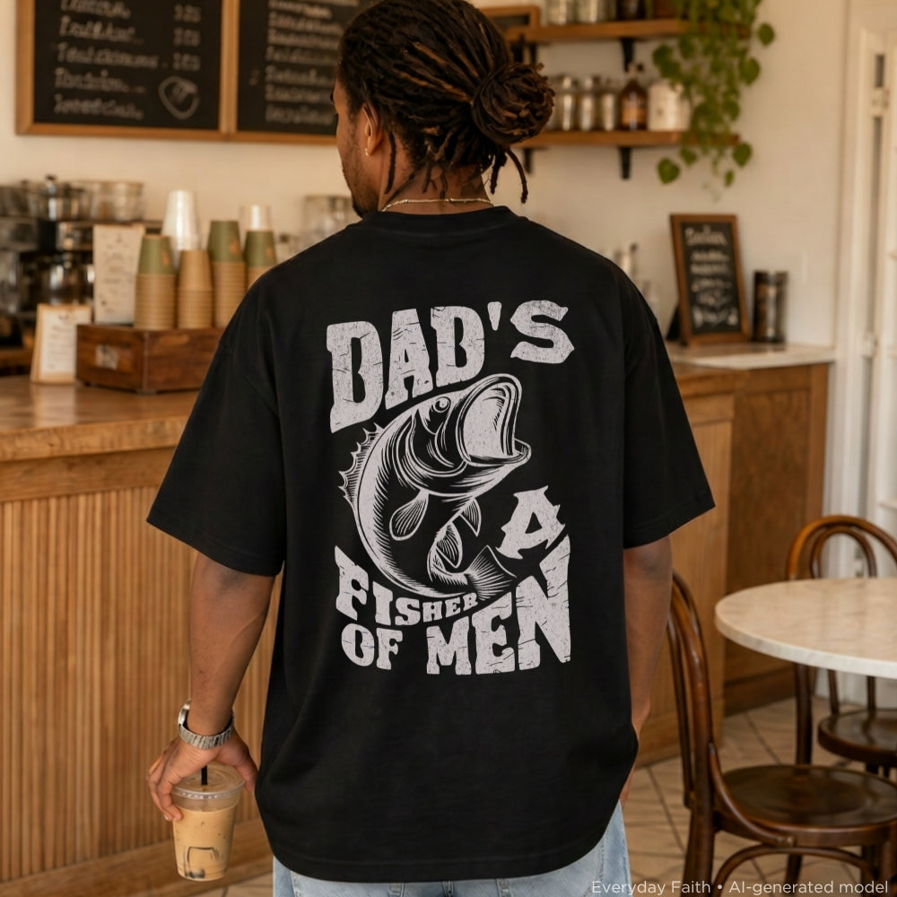 A man viewed from behind wearing a black Everyday Faith t-shirt with a large white graphic that reads 'DAD'S FISHER OF MEN' with a fish illustration, standing in a cafe setting.