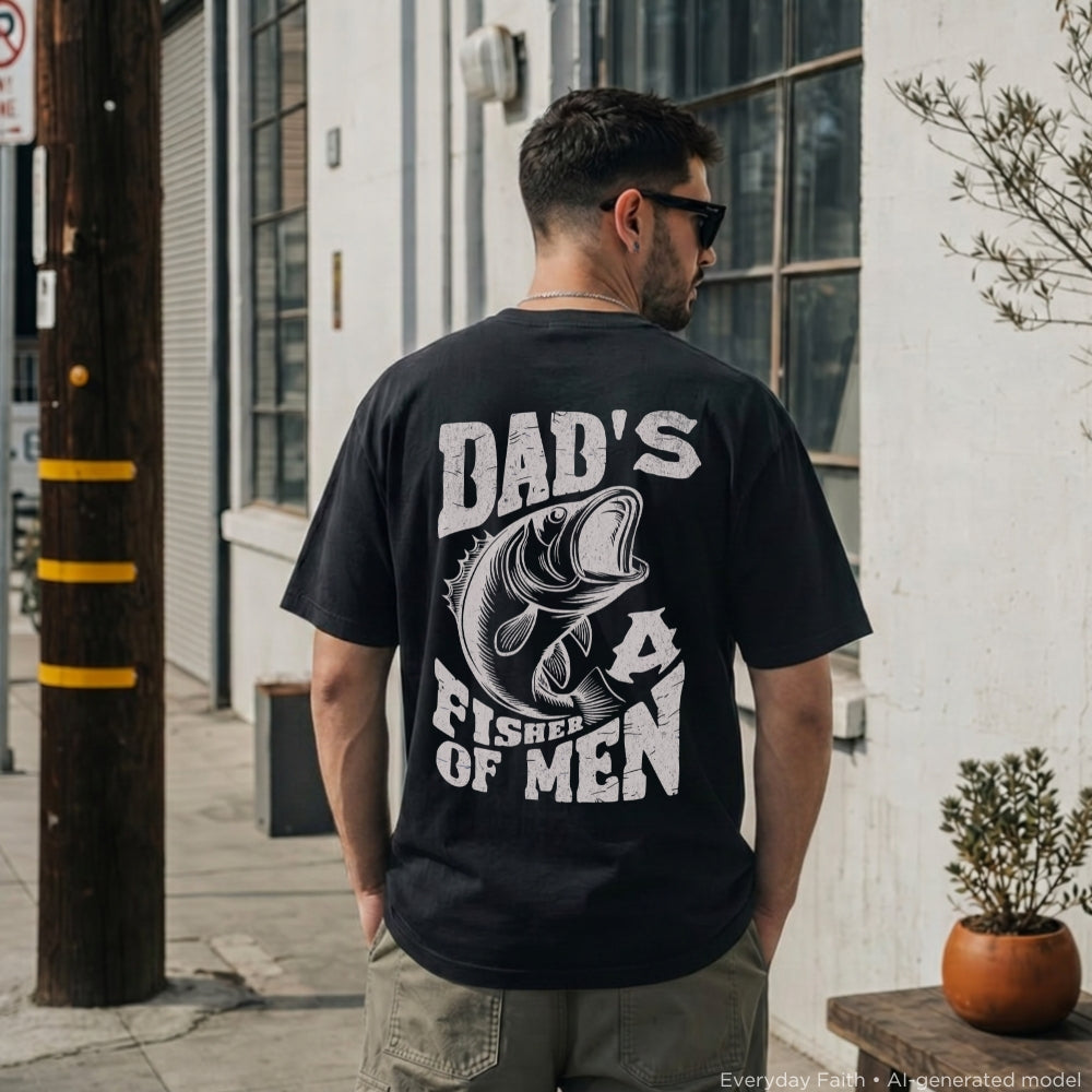 A man wearing a black Everyday Faith graphic t-shirt with a white print that reads 'DAD'S FISHING OF MEN' and features a large bass illustration on the back, standing outdoors on a city sidewalk.