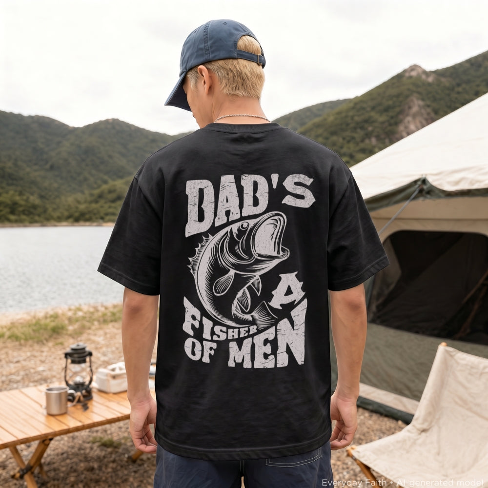 A man viewed from behind wearing a black Everyday Faith t-shirt with a large white graphic that reads 'DAD'S FISHER OF MEN' with a fish illustration, standing outdoors near a lake, mountains, and a tent.