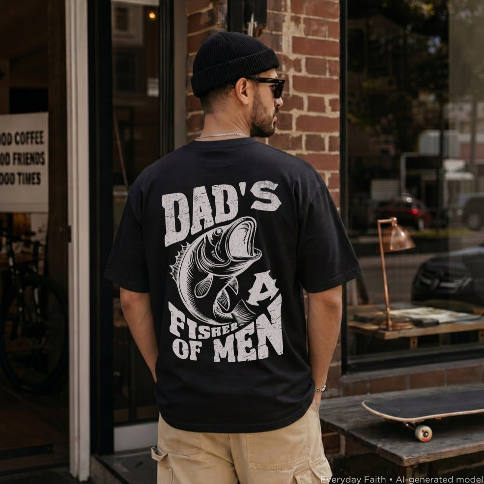 A man wearing a black Everyday Faith t-shirt with a large white graphic that reads 'DAD'S FISHER OF MEN' and features a bass fish illustration, standing in front of a brick storefront.
