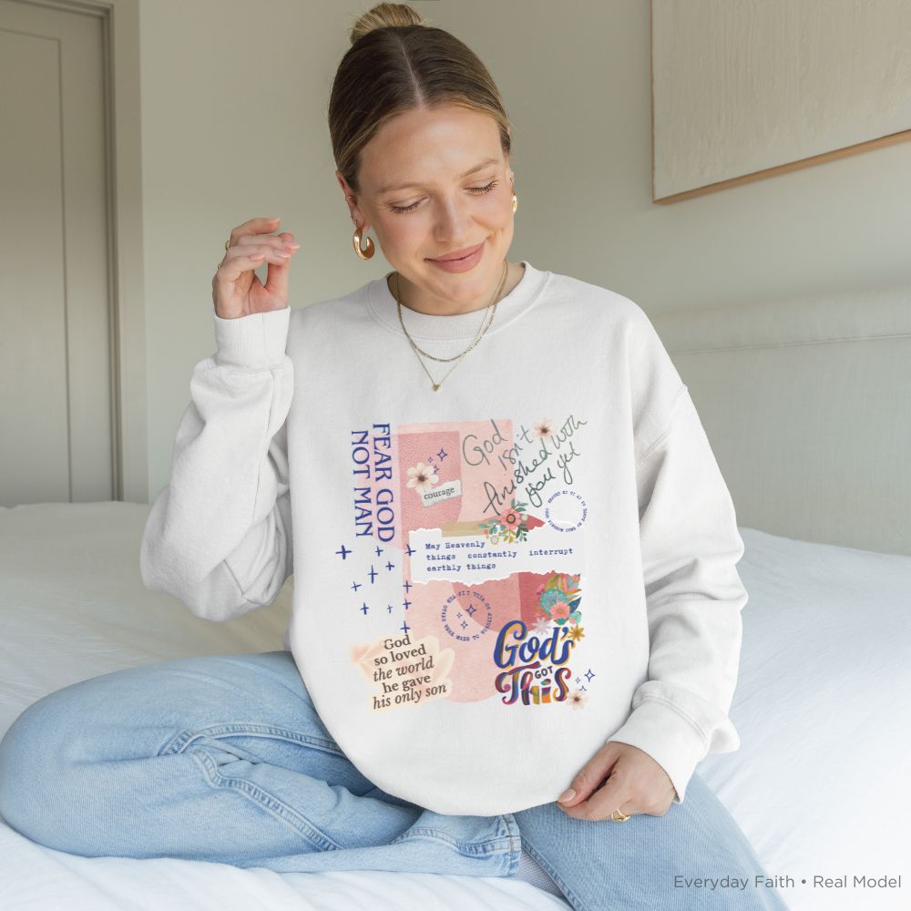 A woman sitting on a bed wearing a white Everyday Faith crewneck sweatshirt with a colourful graphic print that reads 'Fear God Not Man' and 'God is Love, Love is Enough' with floral and script details. She is wearing light blue jeans.