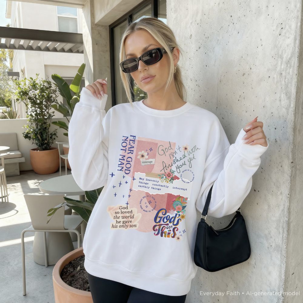 A woman wearing a white Everyday Faith crewneck sweatshirt with a colourful graphic collage of religious quotes and illustrations, styled with black sunglasses and a black shoulder bag, leaning against a concrete wall in an outdoor patio setting.
