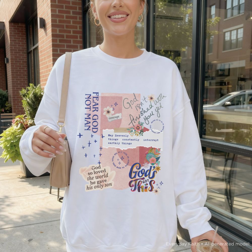 A woman wearing a white long-sleeve Everyday Faith crewneck sweatshirt with a colourful graphic collage featuring religious quotes, flowers, and doodles. She is standing outdoors in front of a brick building and glass storefront.