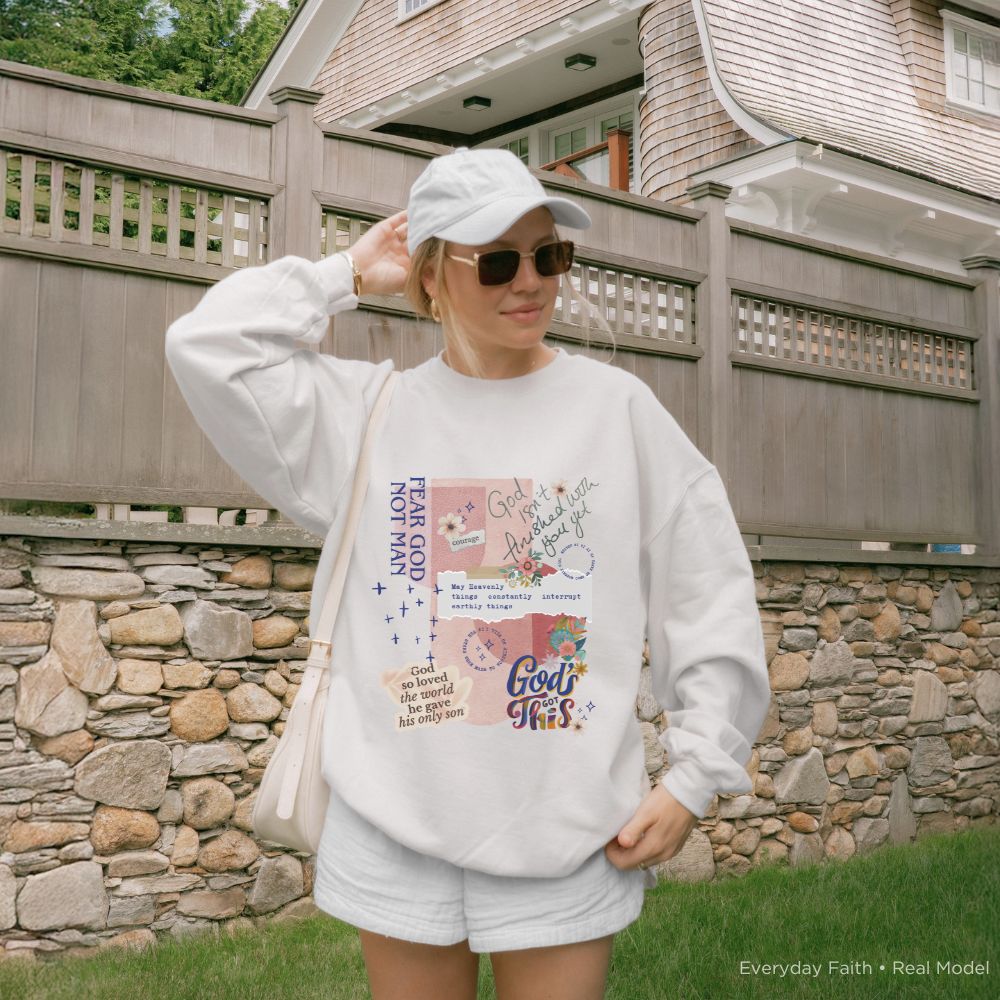 A woman wearing a white Everyday Faith graphic sweatshirt and matching white shorts stands outdoors in front of a stone wall and wooden fence. She is also wearing a white cap, sunglasses, and carrying a light-coloured shoulder bag.