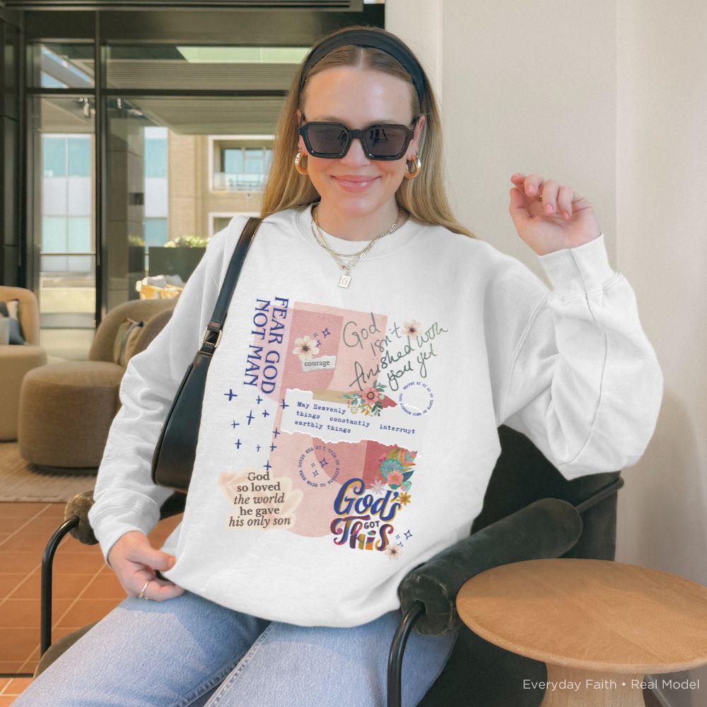 A woman wearing a white Everyday Faith graphic sweatshirt with inspirational text and floral collage designs, sitting in a modern indoor setting.