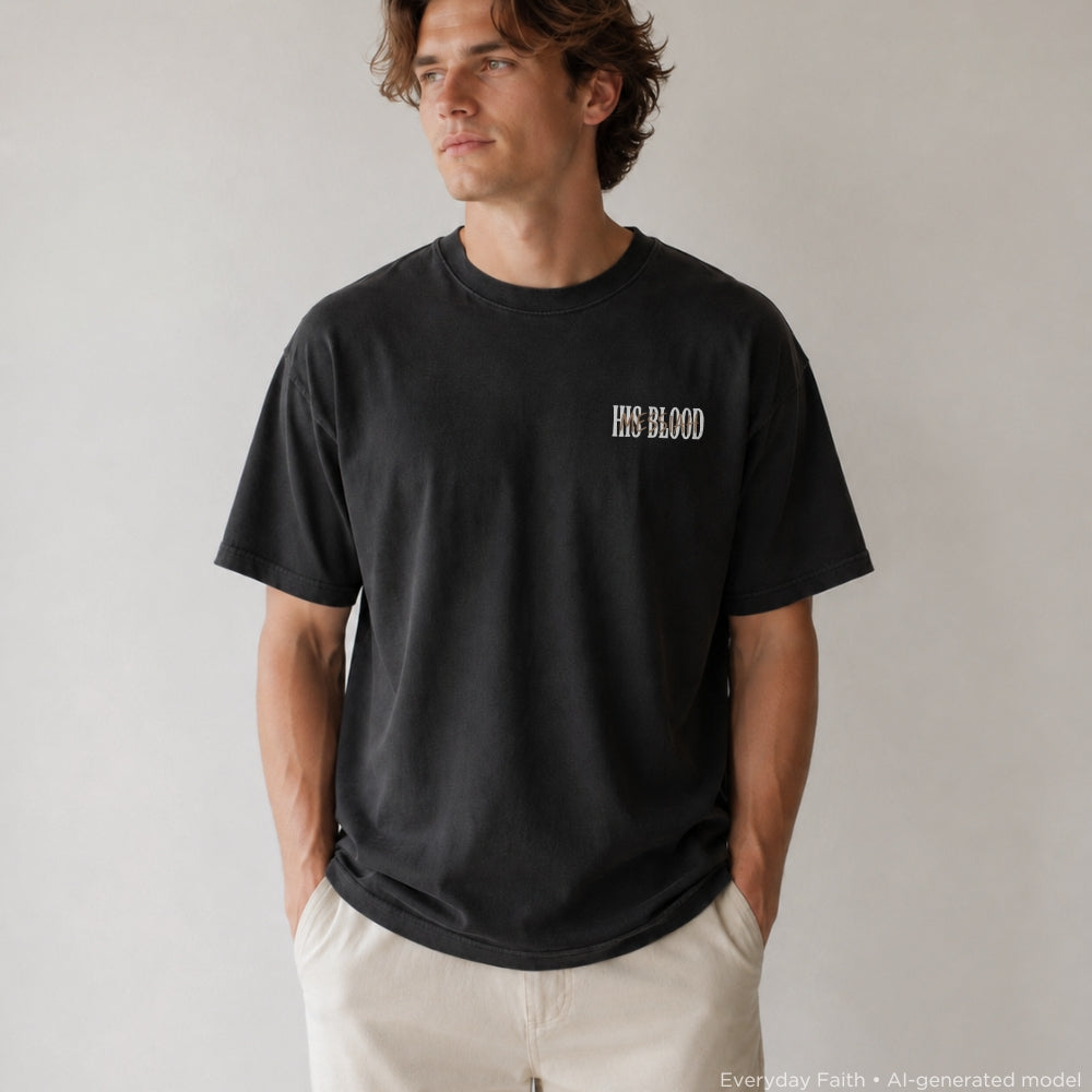 A man wearing a black Everyday Faith crewneck t-shirt with 'HIS BLOOD' printed in white on the left chest, paired with off-white pants, standing against a neutral light gray background.