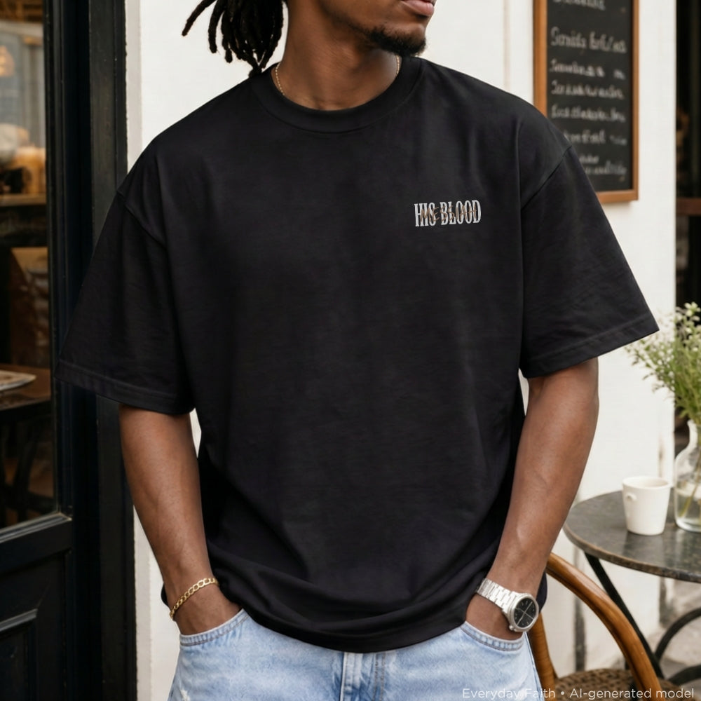 A man wearing a black Everyday Faith oversized t-shirt with 'HIS BLOOD' printed in white on the left chest, paired with light blue jeans, standing in front of a cafe storefront.