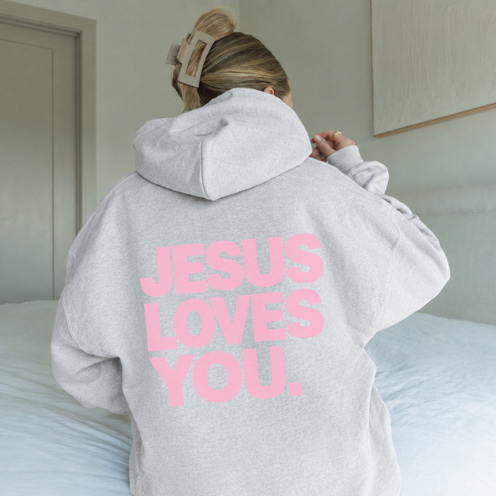 A person seen from behind wearing a light gray oversized Everyday Faith hoodie with the phrase 'JESUS LOVES YOU.' printed in large pink letters on the back, sitting on a bed in a bedroom.