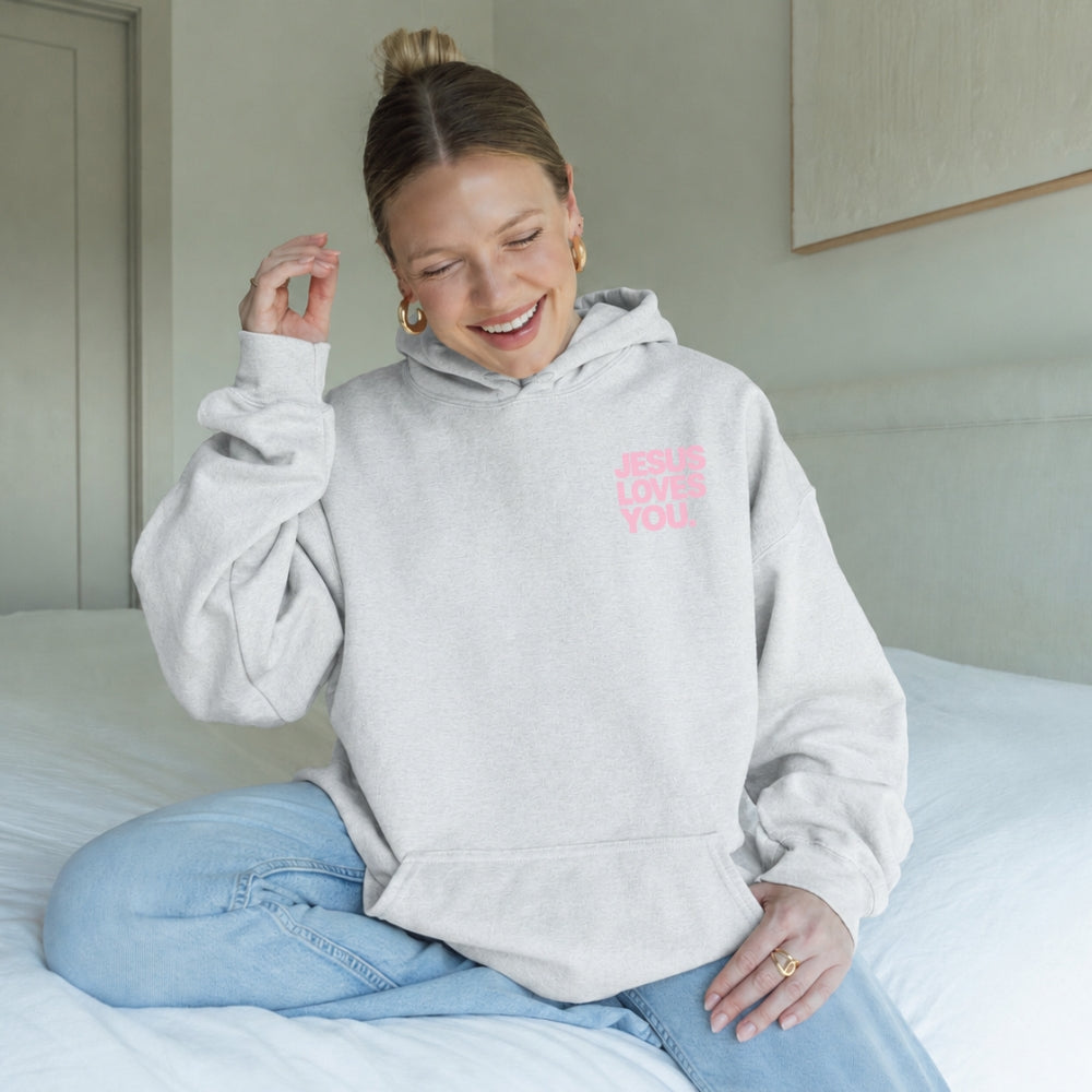 A person sitting on a bed wearing a light gray Everyday Faith hoodie with pink text that reads 'JESUS LOVES YOU' on the left chest, paired with light blue jeans.