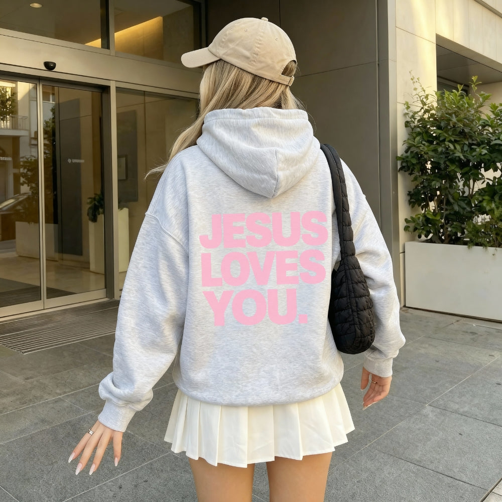 A person viewed from behind wearing a light gray heathered Everyday Faith hoodie with 'JESUS LOVES YOU.' printed in pink across the back, paired with a cream pleated mini skirt, a beige baseball cap, and a black quilted shoulder bag, standing outside a modern building.