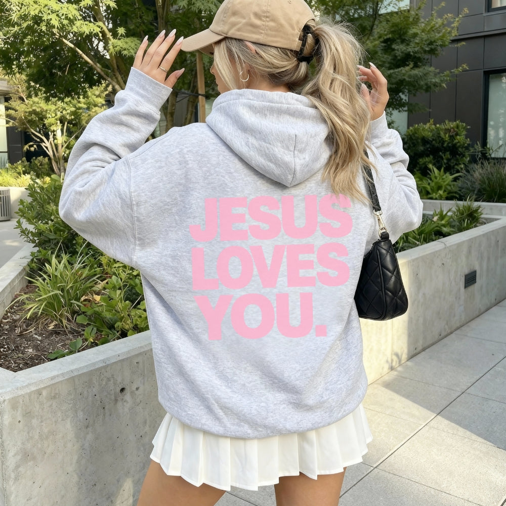 A person wearing a light heather gray Everyday Faith hoodie with the phrase 'JESUS LOVES YOU.' printed in bold pink letters across the back, paired with a white pleated skirt and a tan baseball cap, photographed outdoors on a paved walkway with greenery and a building in the background.