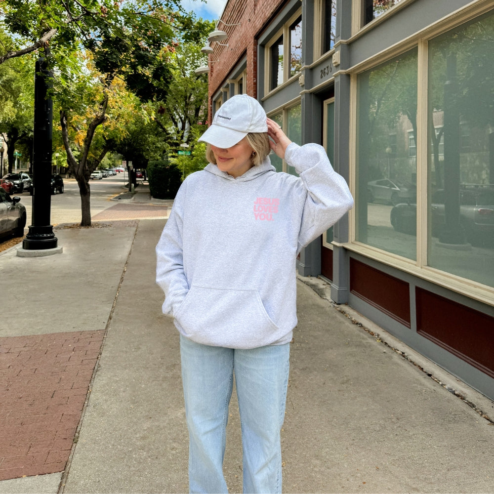 A person stands on a city sidewalk wearing a light gray Everyday Faith hoodie with a small pink heart graphic on the chest, light blue jeans, and a white baseball cap.