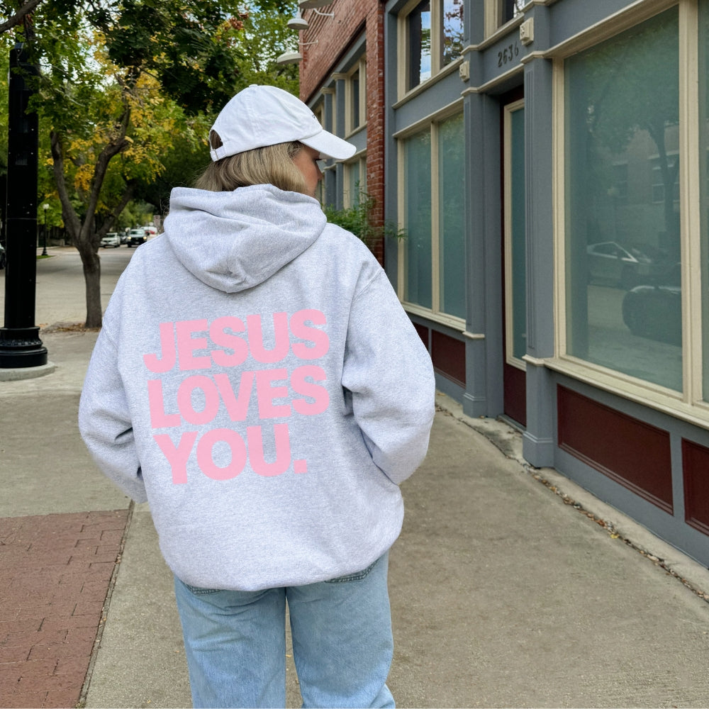 A person standing on a city sidewalk, seen from behind, wearing a light gray Everyday Faith hoodie with large pink text reading 'JESUS LOVES YOU.' on the back, a white baseball cap, and light blue jeans.
